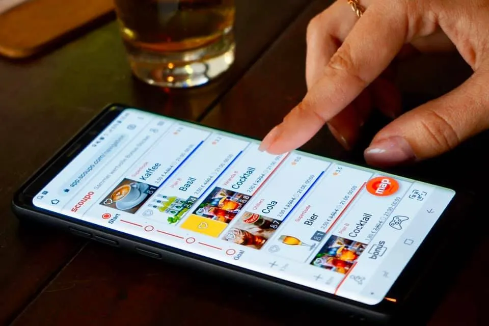 Person bedient die Scoupo-App auf einem Smartphone in einer Bar, im Vordergrund ein Glas Bier und eine Hand, die auf einen Cocktail-Coupon tippt.